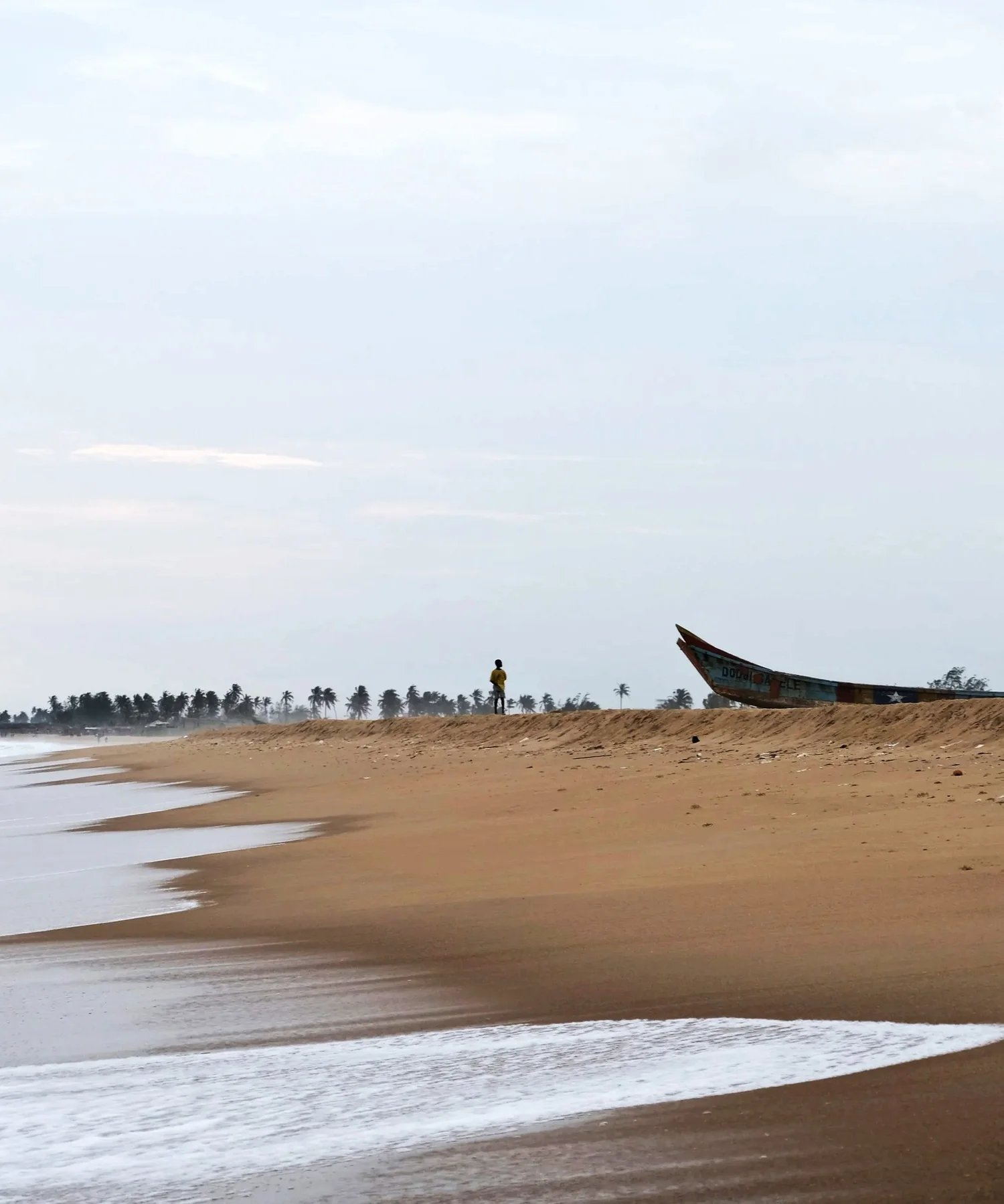 A person standing on a sandy beach near the shoreline, with a colorful boat on the sand nearby and palm trees in the distance under a cloudy sky.