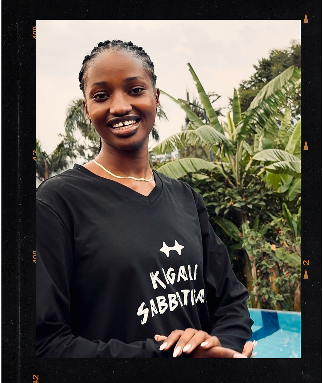 A young woman smiling outdoors, wearing a black shirt with the text 'KIGALI SABBITO' and a small logo, with lush green plants in the background.
