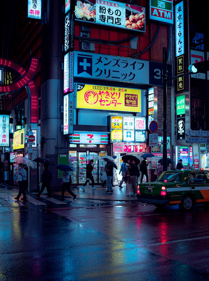 Nighttime street scene in a busy district with illuminated signs, pedestrians with umbrellas crossing wet streets, and a green and white taxi cab.