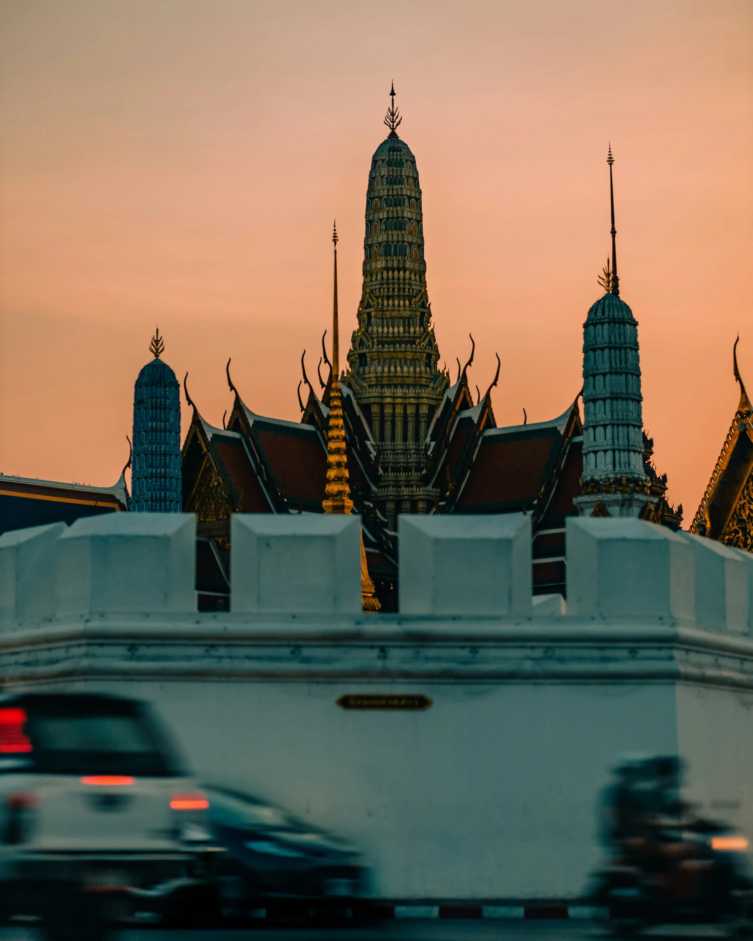 A temple with traditional Thai architecture seen at sunset, with cars passing by in the foreground.