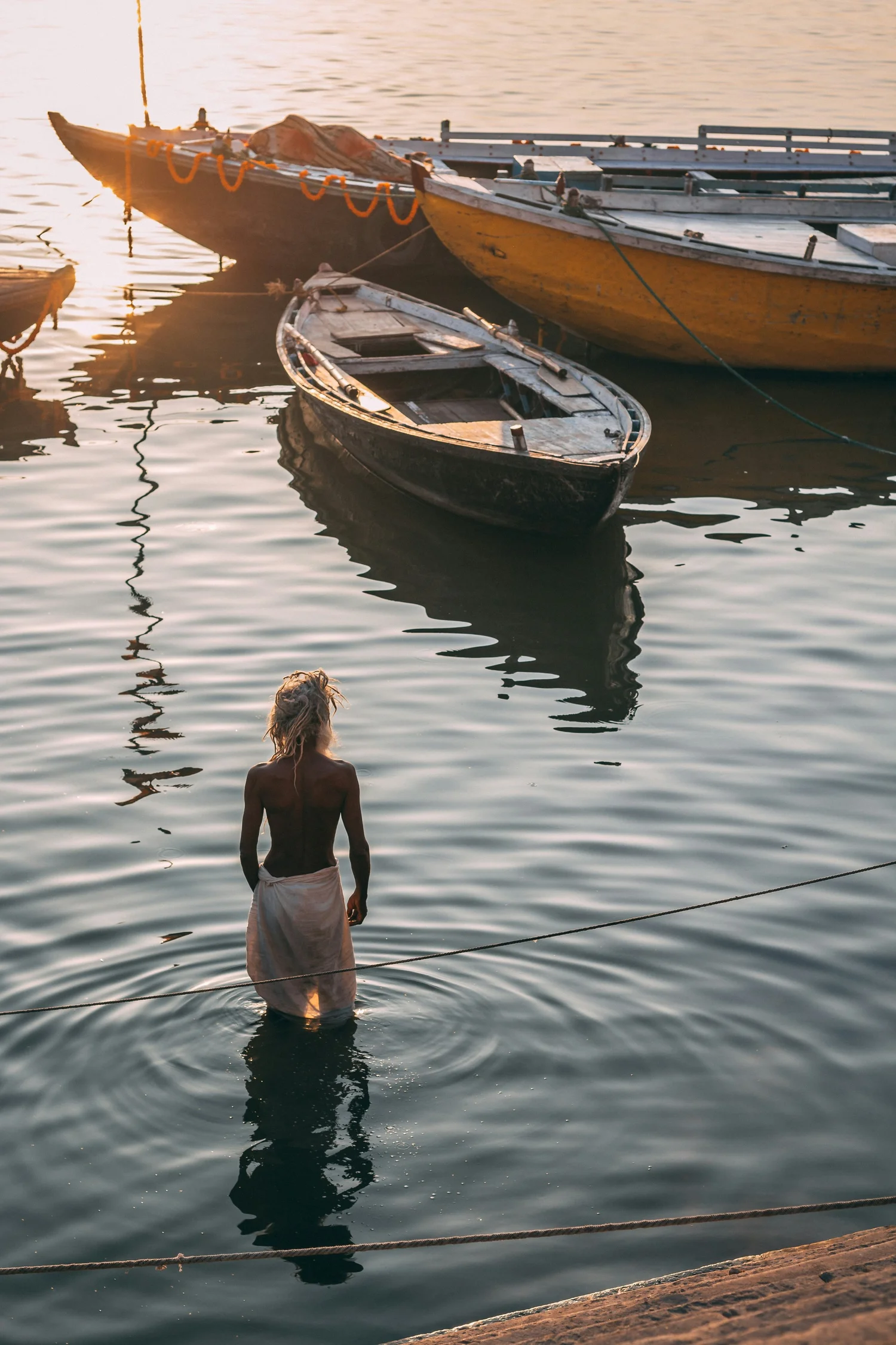 A shirtless man with long hair, wearing light-colored shorts, wades in the water near a dock with moored boats, during sunset.