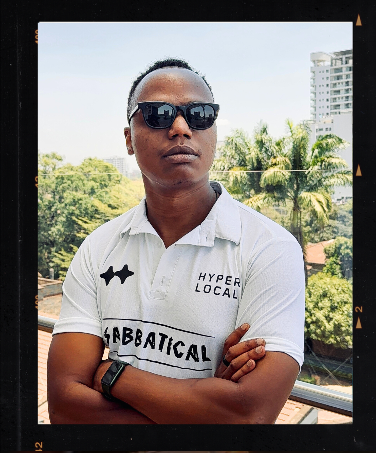 A man wearing sunglasses and a white polo shirt with black print, standing outdoors with arms crossed, high-rise buildings, trees, and a blue sky in the background.