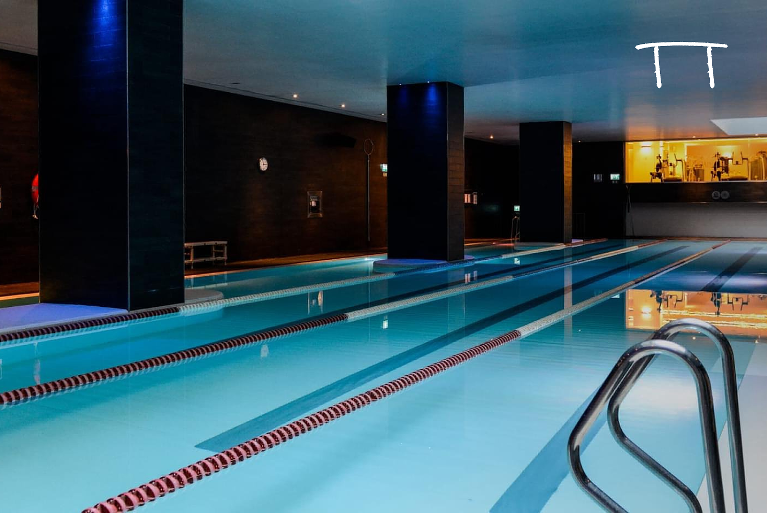 Indoor swimming pool with multiple lanes, dark walls, and a reflection of the ceiling lights on the water surface.