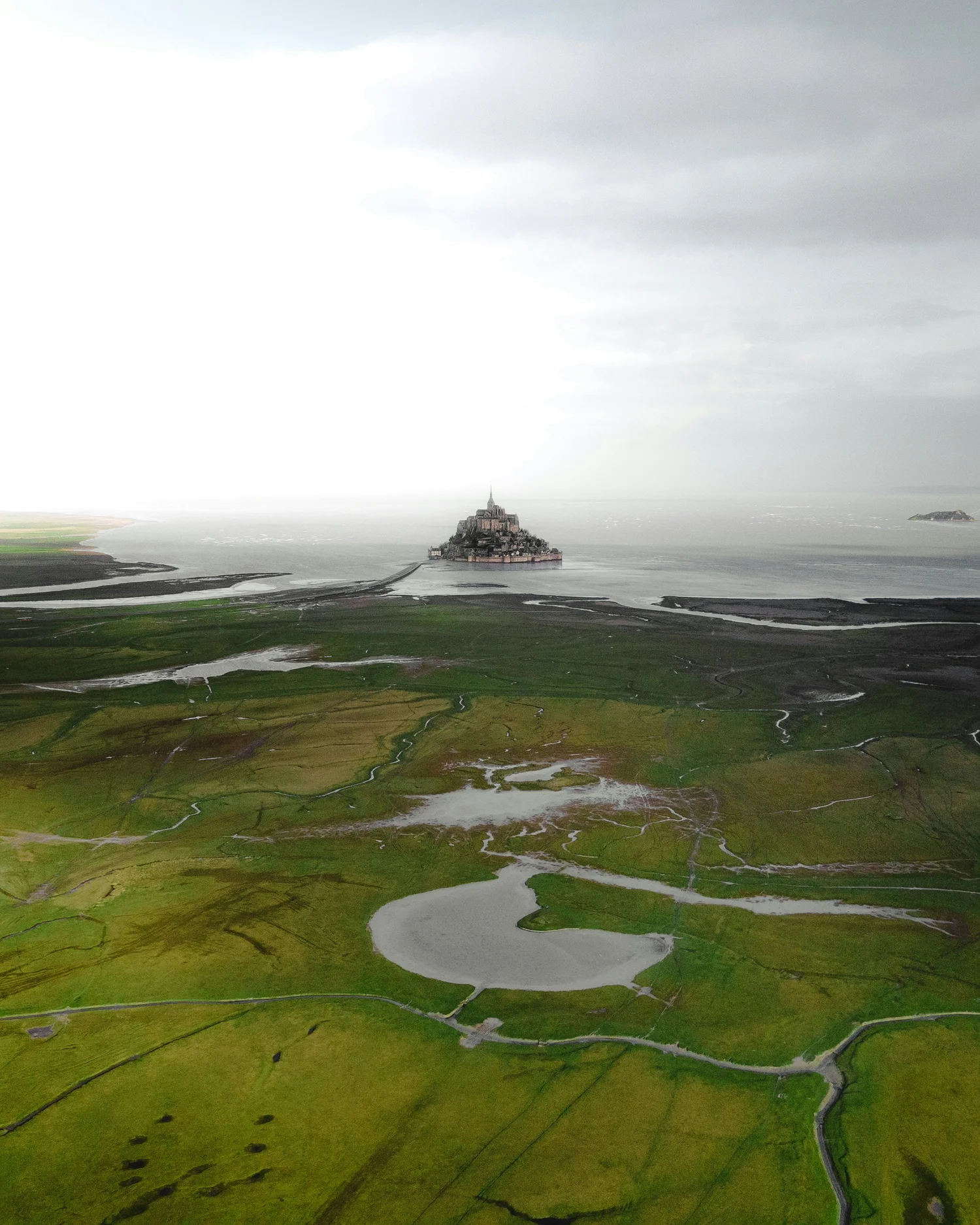 Aerial view of Mont Saint-Michel, a historic island commune in Normandy, France, surrounded by green fields, waters, and connected to the mainland by a causeway under an overcast sky.