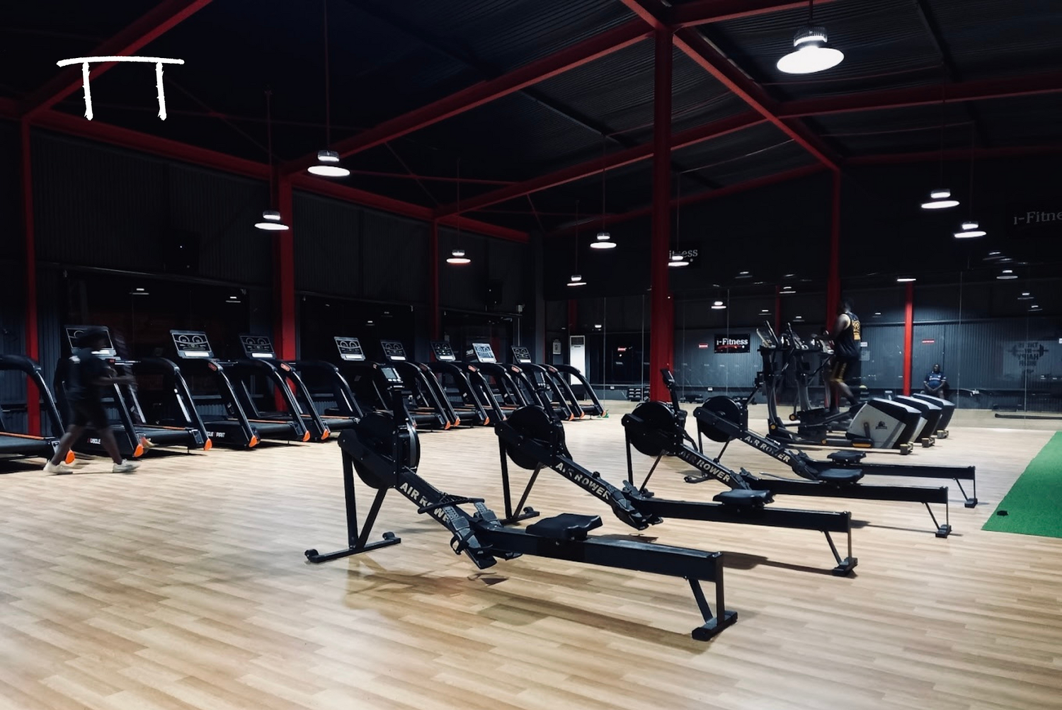 Empty gym with treadmills, rowing machines, and a person running on a treadmill. The gym has wooden floors, red and black walls, and bright ceiling lights.