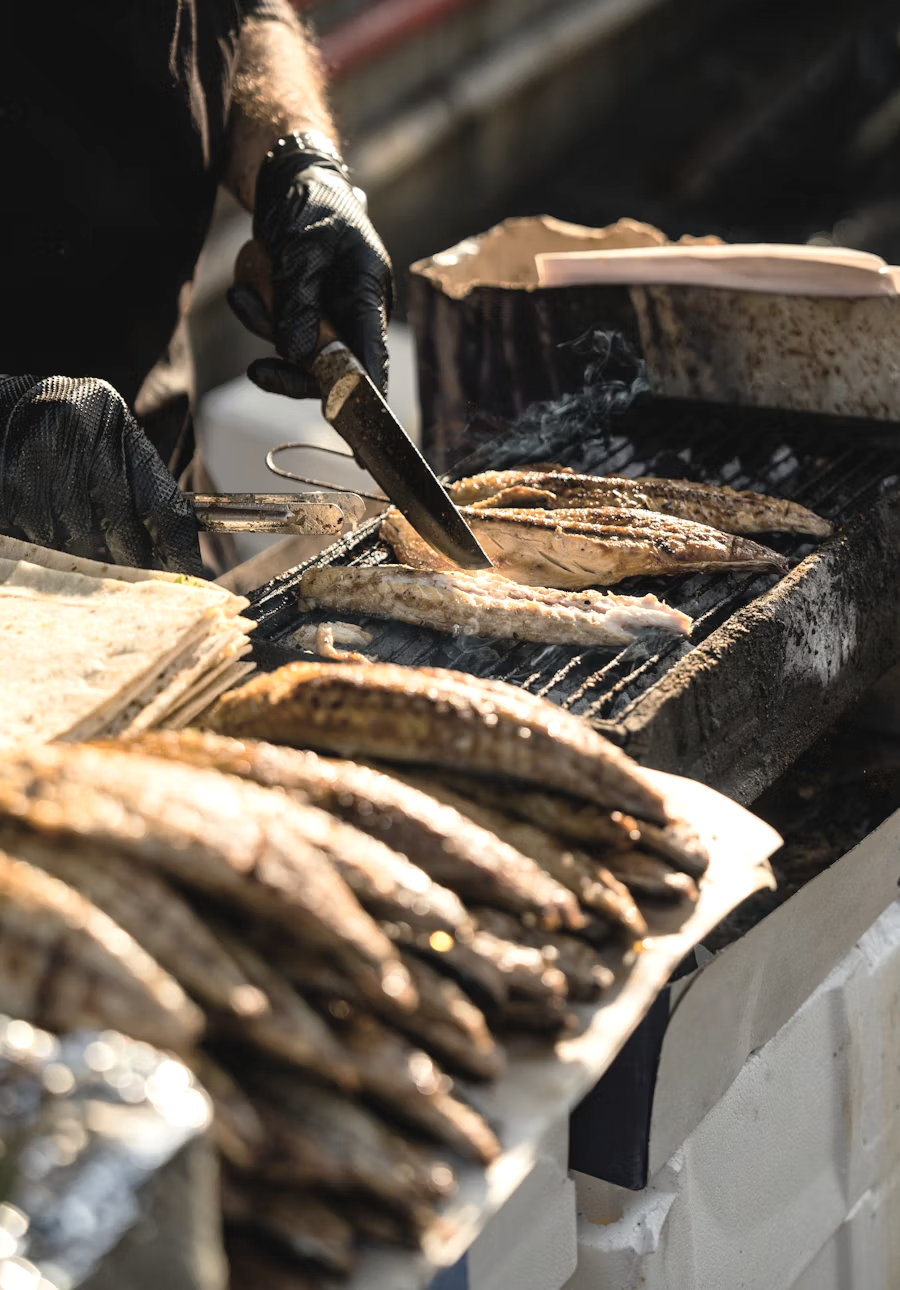 Person wearing black gloves cooking multiple fish on a grill outdoors.