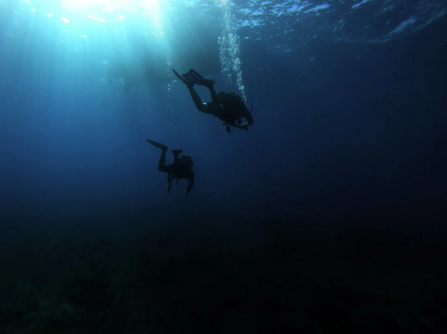 Two scuba divers exploring underwater, with sunlight shining down from above and dark water below.