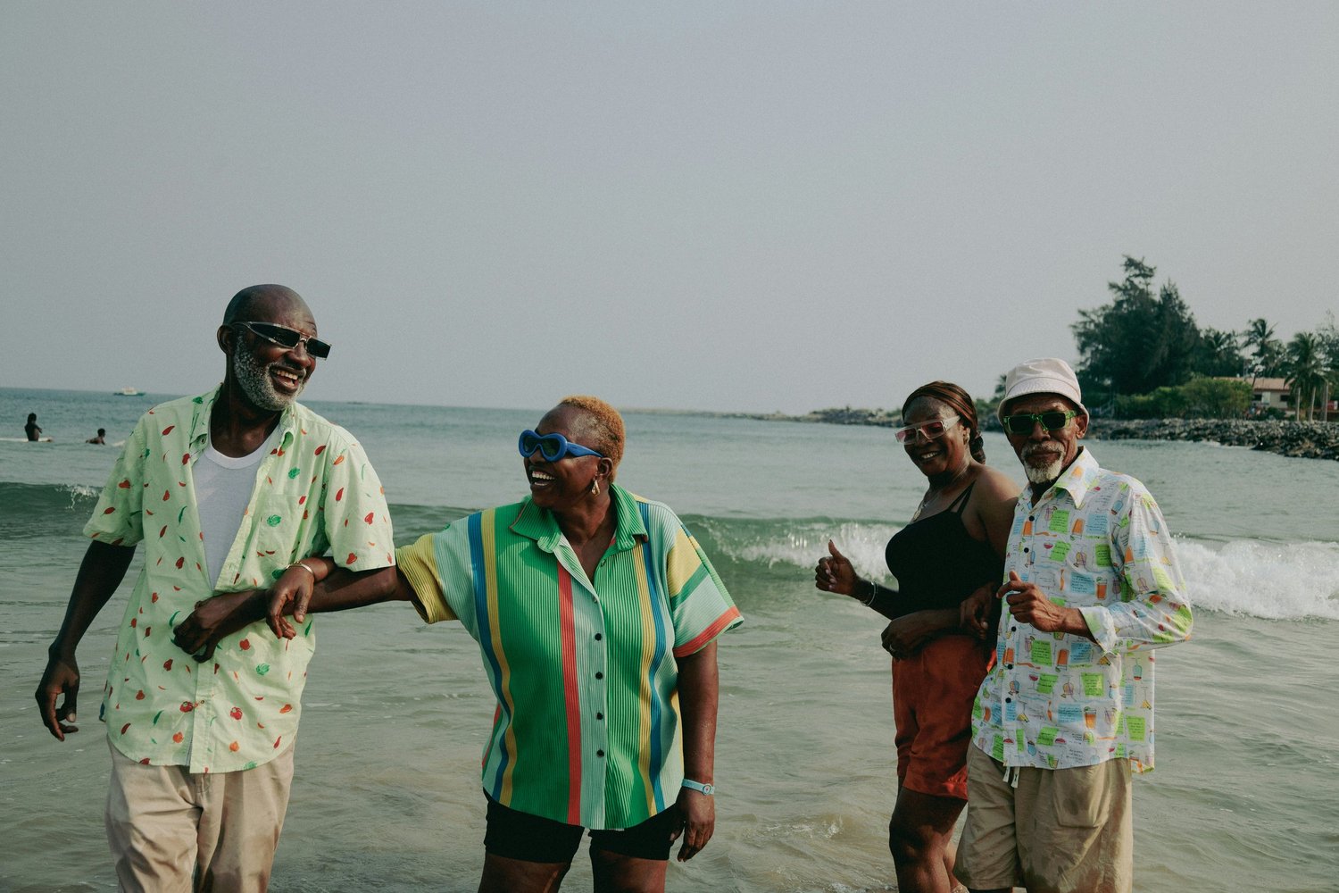 Four elderly people dressed in colorful summer clothes standing in the ocean waves at the beach, smiling and enjoying the time together.