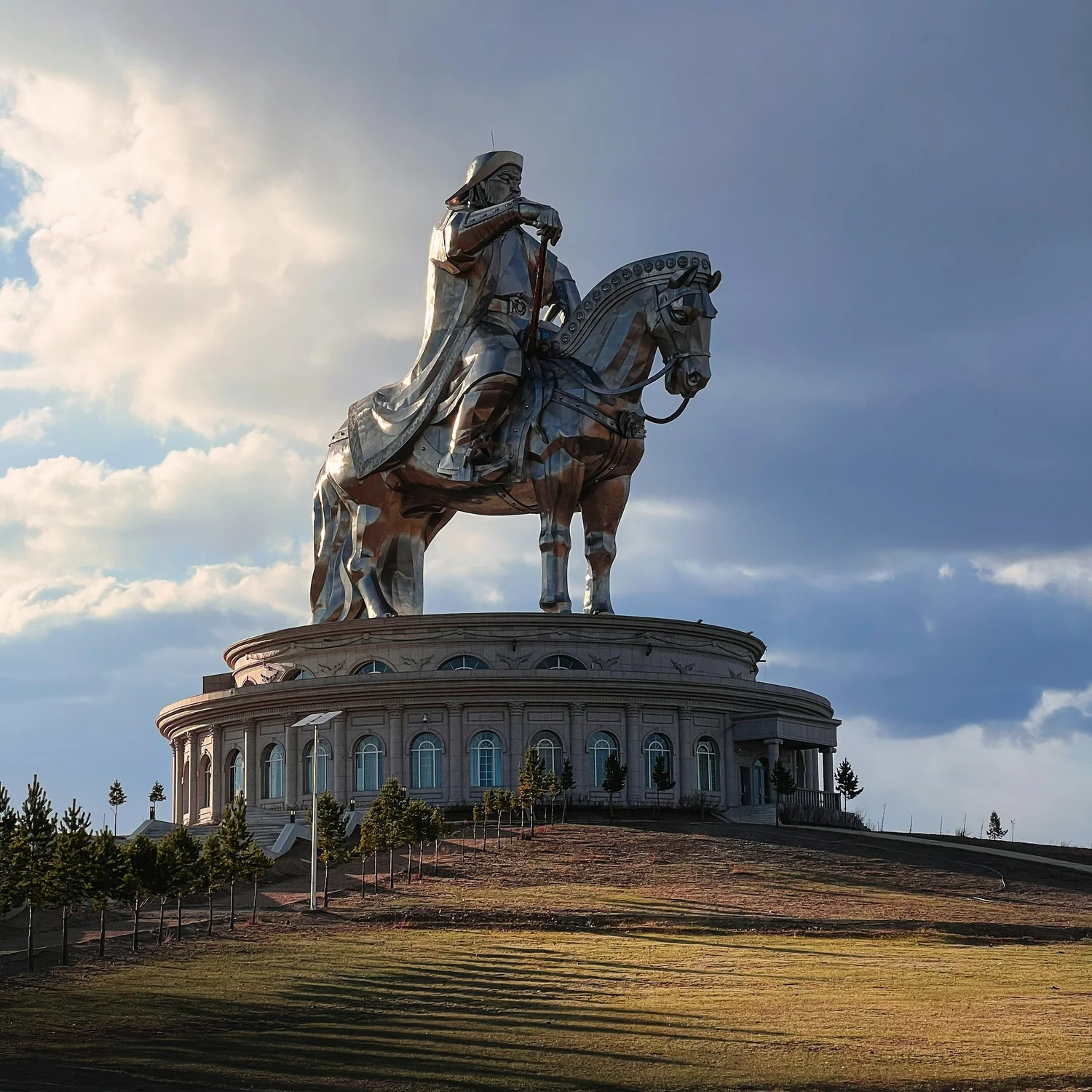 A large silver equestrian statue of a man on a horse, located on a circular stone platform with small windows and surrounded by small trees, under a partly cloudy sky.