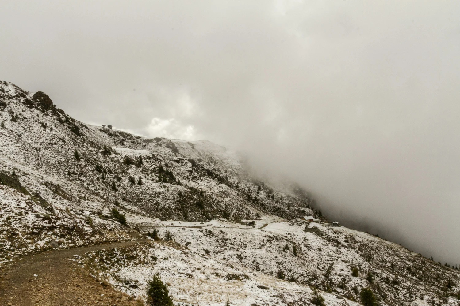 Snow-covered mountain landscape with a dirt road, scattered trees, and foggy clouds in the sky.