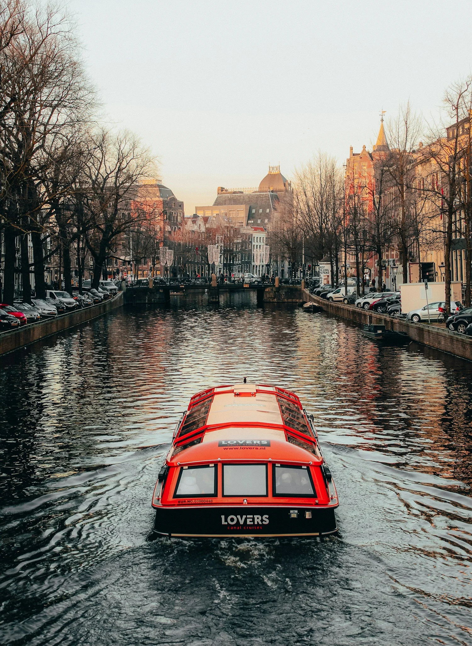 A red canal cruise boat on a canal in a European city, with historic buildings and leafless trees on either side during winter.