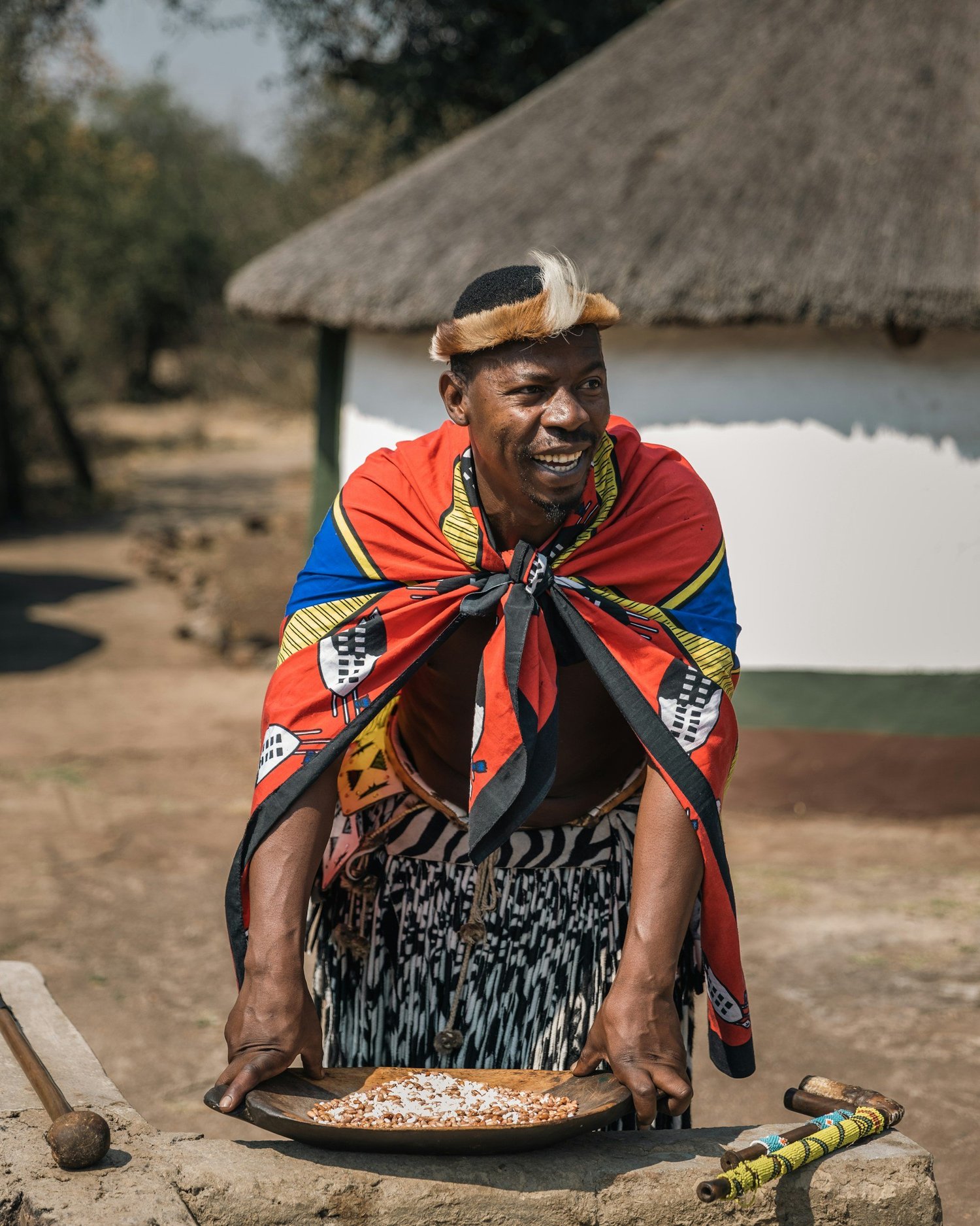 A smiling man dressed in traditional African attire, draped in a brightly colored cloth, leaning forward on a table with food and musical instruments outdoors.