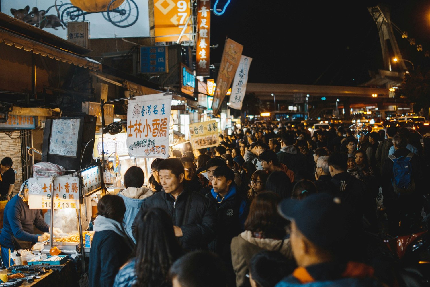 Night market scene packed with people, food stalls, colorful signs in Chinese, and bright lights.