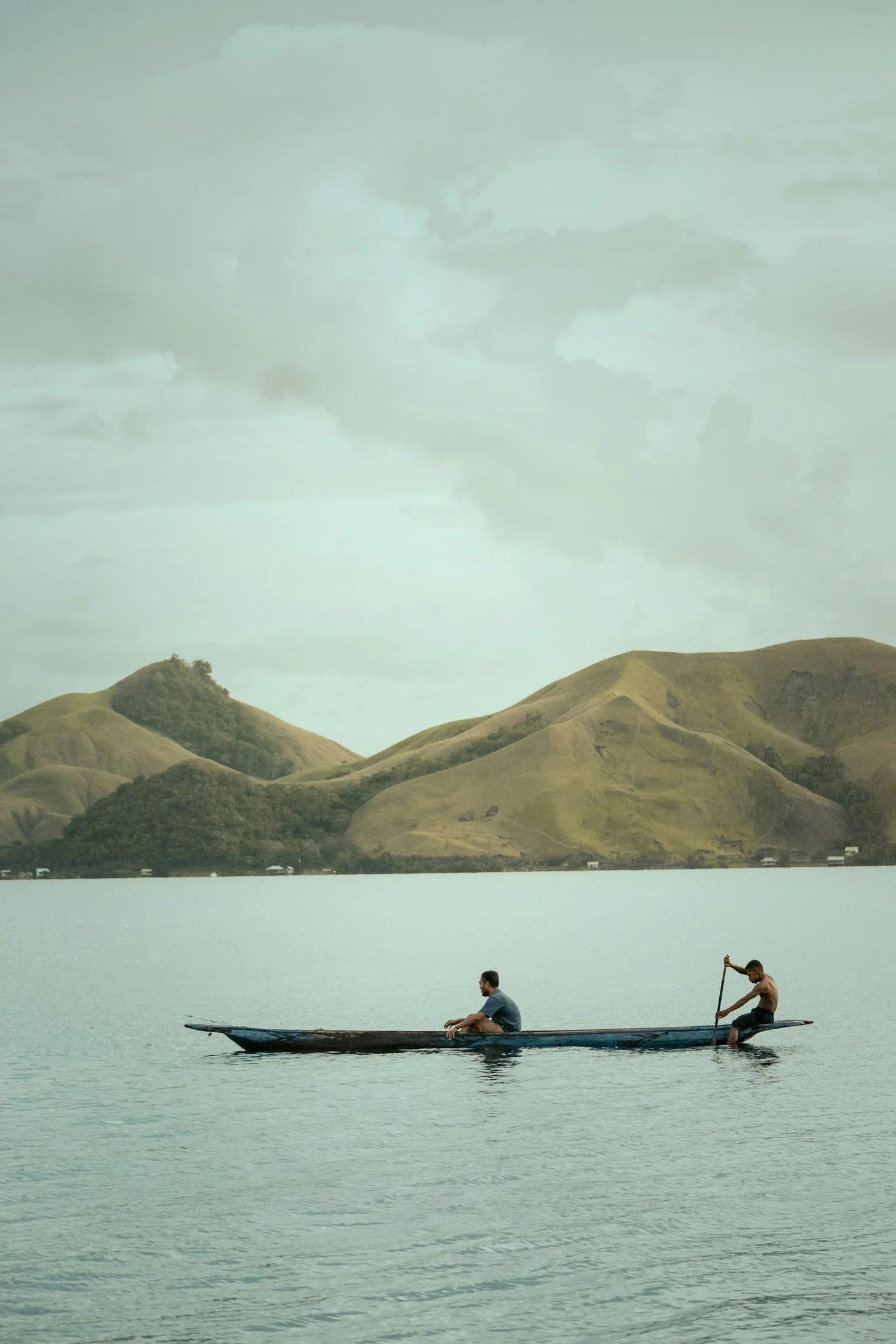 Two men in a canoe on a calm body of water with green hills in the background and cloudy sky above.