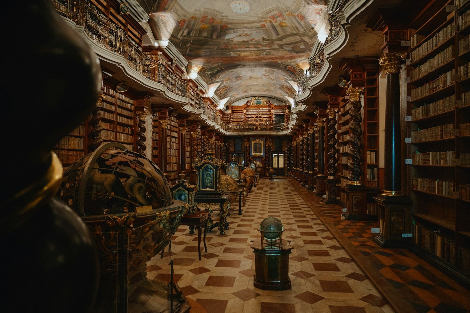 An ornate historic library with tall wooden shelves filled with books, decorative columns, and a painted ceiling. There are globes and antique artifacts displayed along the aisle.