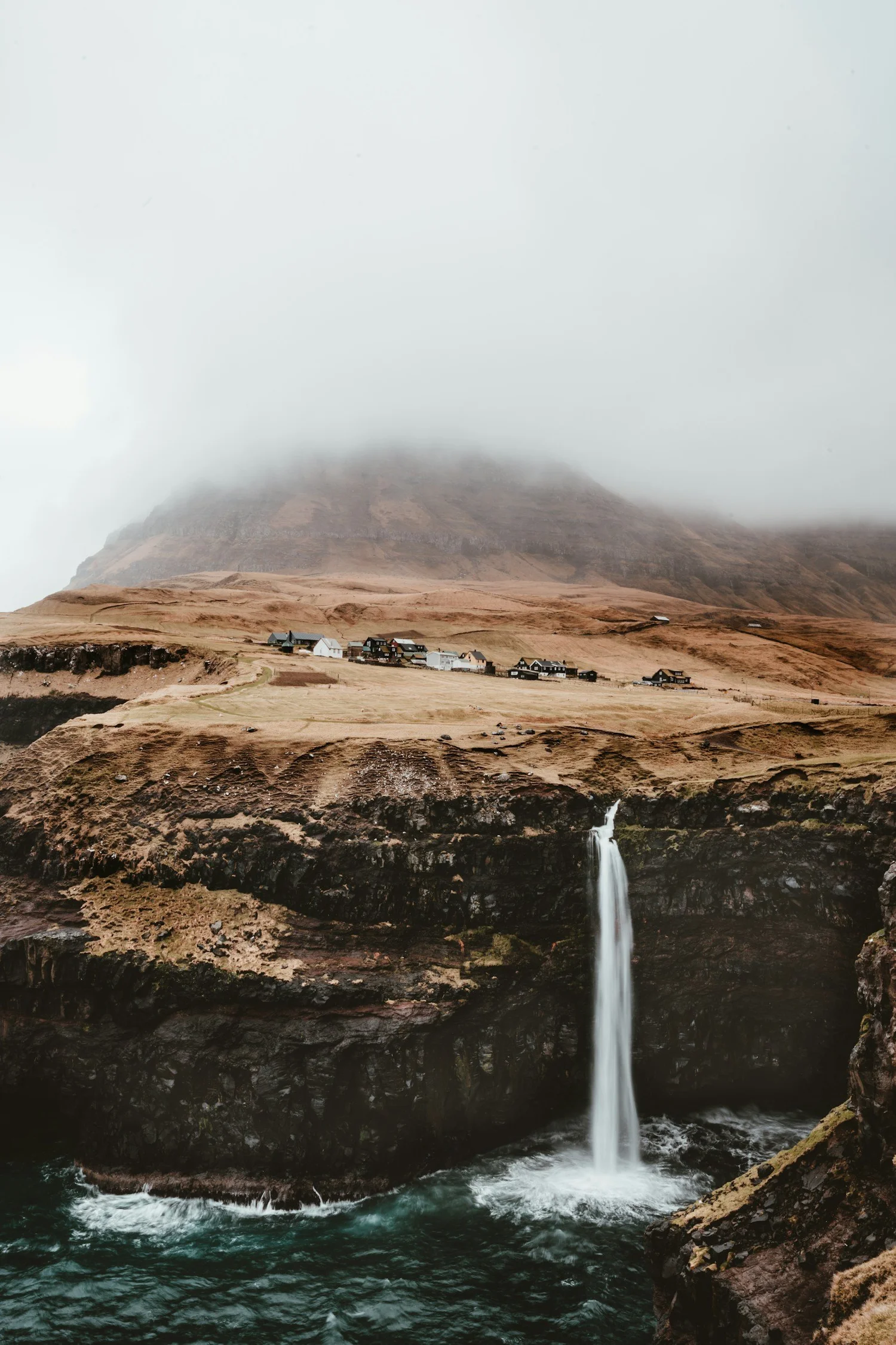 A landscape with a waterfall cascading into a dark blue river, with a small village of houses on a brown grassy hillside beneath a fog-covered mountain.