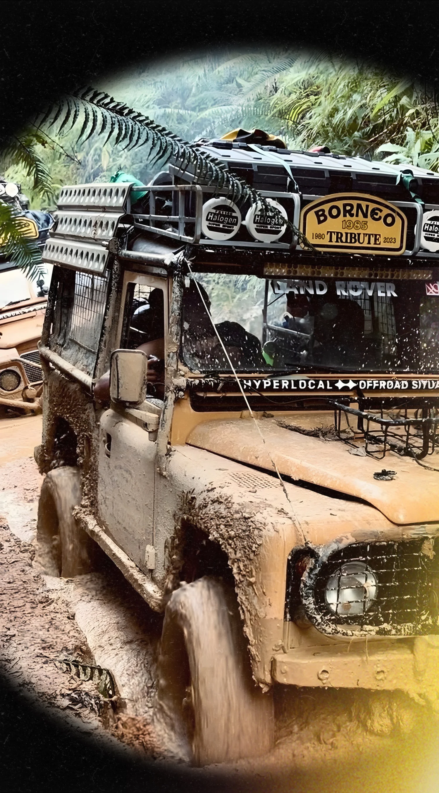 A muddy off-road safari vehicle splashing mud as it drives through a muddy terrain, with jungle foliage in the background and a sign that reads 'Bornéo Tribute 1985-2023'.