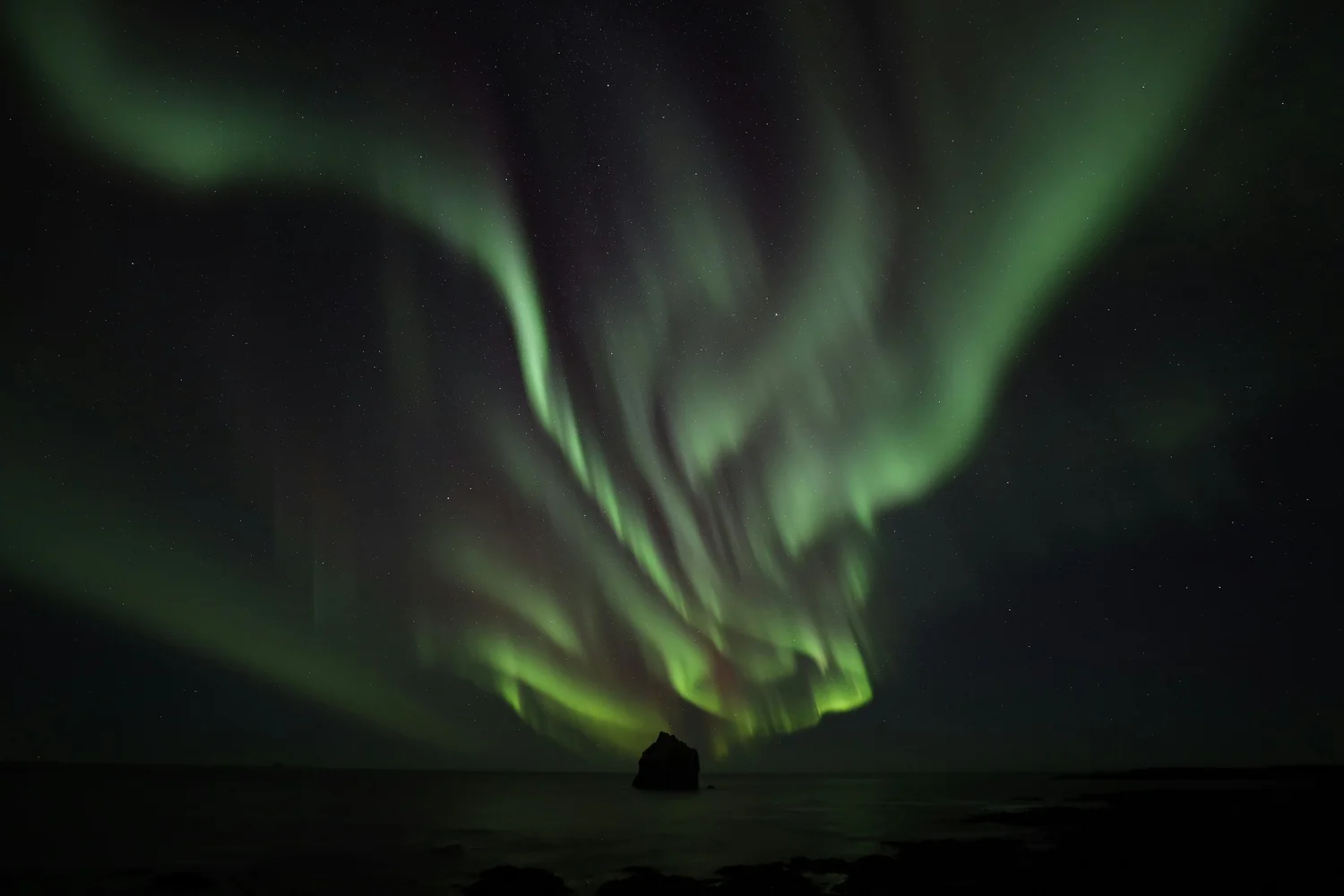 Northern lights over a dark ocean with a large rock near the shoreline.