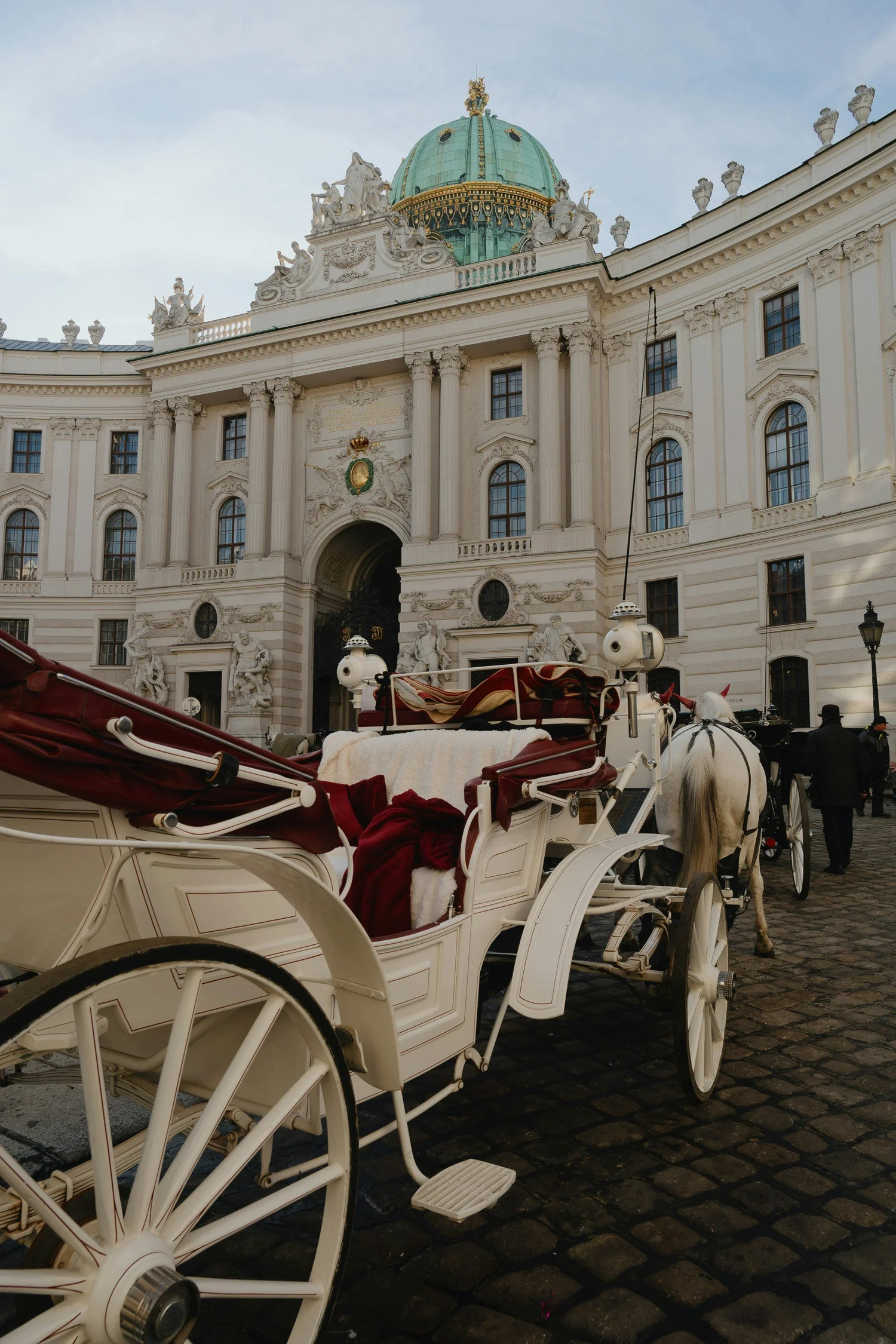 A horse-drawn carriage parked in front of a grand, historic building with classical architectural details, including a green dome, sculptures, and ornate decorations, on a cobblestone street.
