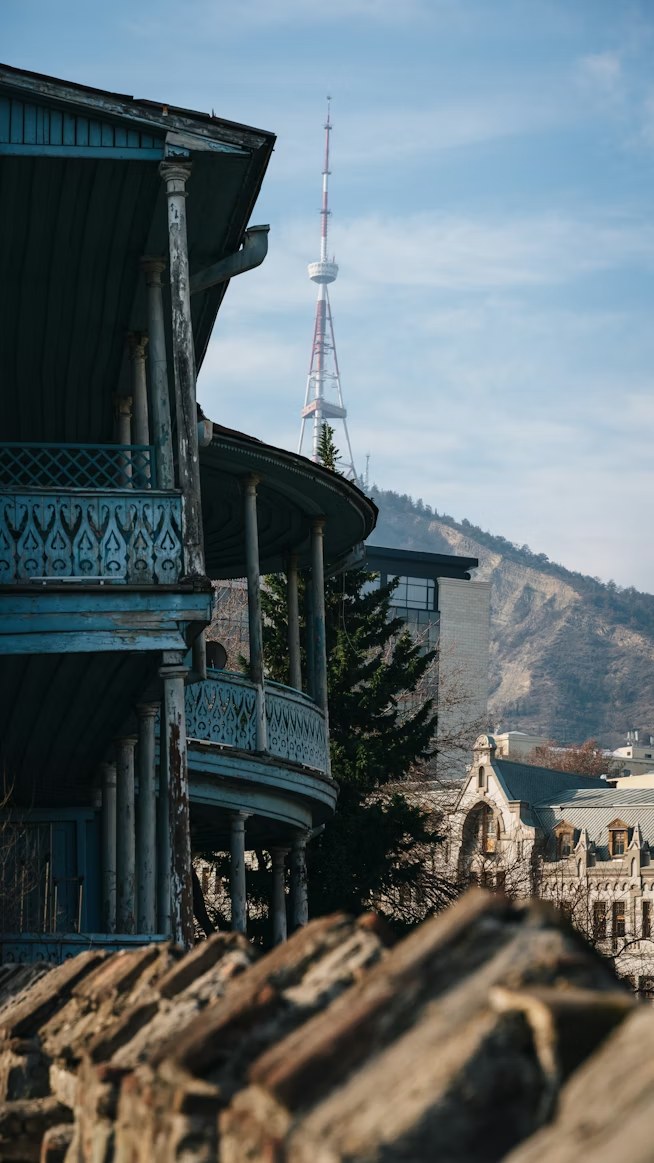 A vintage blue house with ornate balcony railings and peeling paint, overlooking a cityscape with a mountain and a tall communications tower in the background.