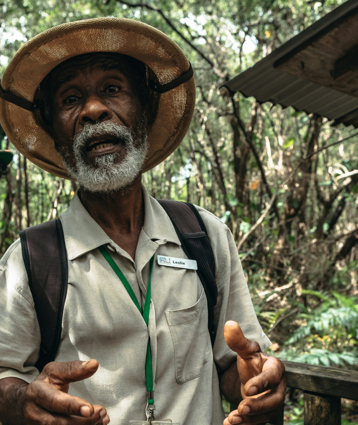 An older man with a white beard and gray hair, wearing a straw hat with a black band, a beige shirt with a name tag that says 'Leslie,' and a green lanyard, standing in a lush forest. He is carrying a backpack and gesturing with his hands while speaking.