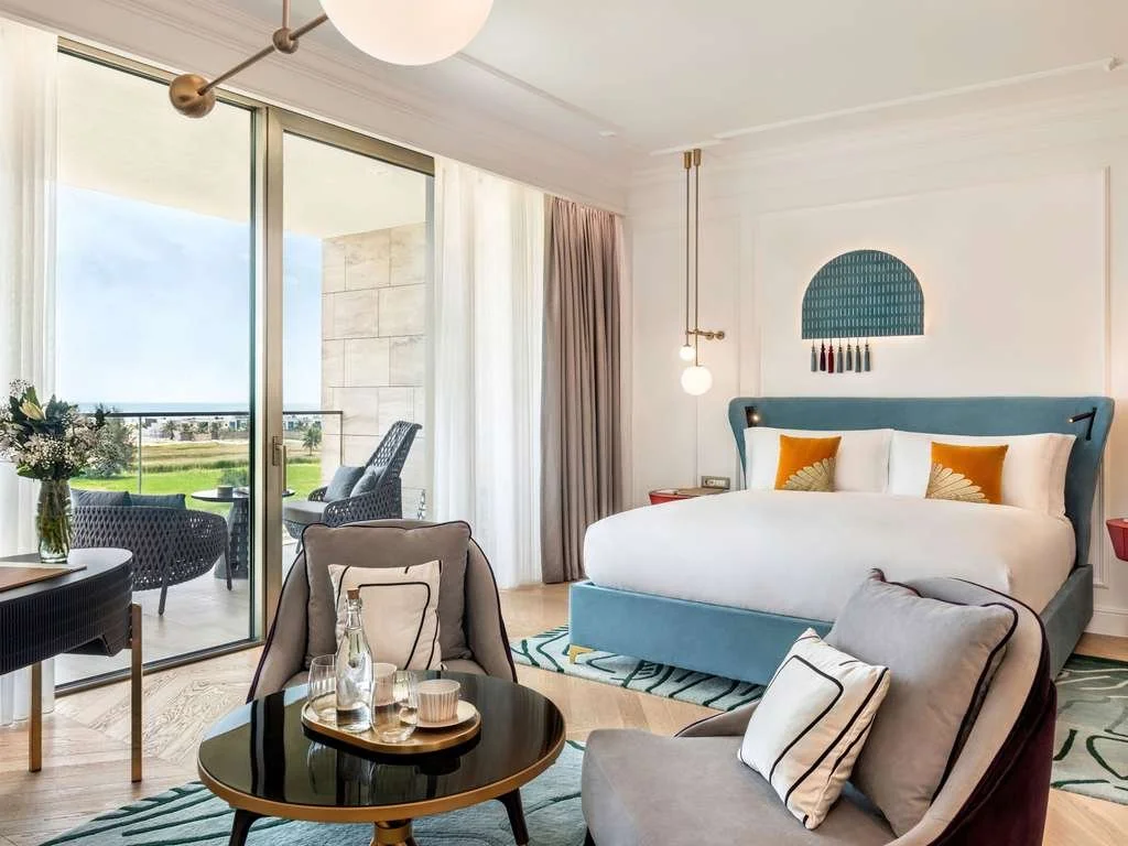 A modern hotel room with a large bed, a small table with water bottles, and a balcony with outdoor seating and a view of greenery.
