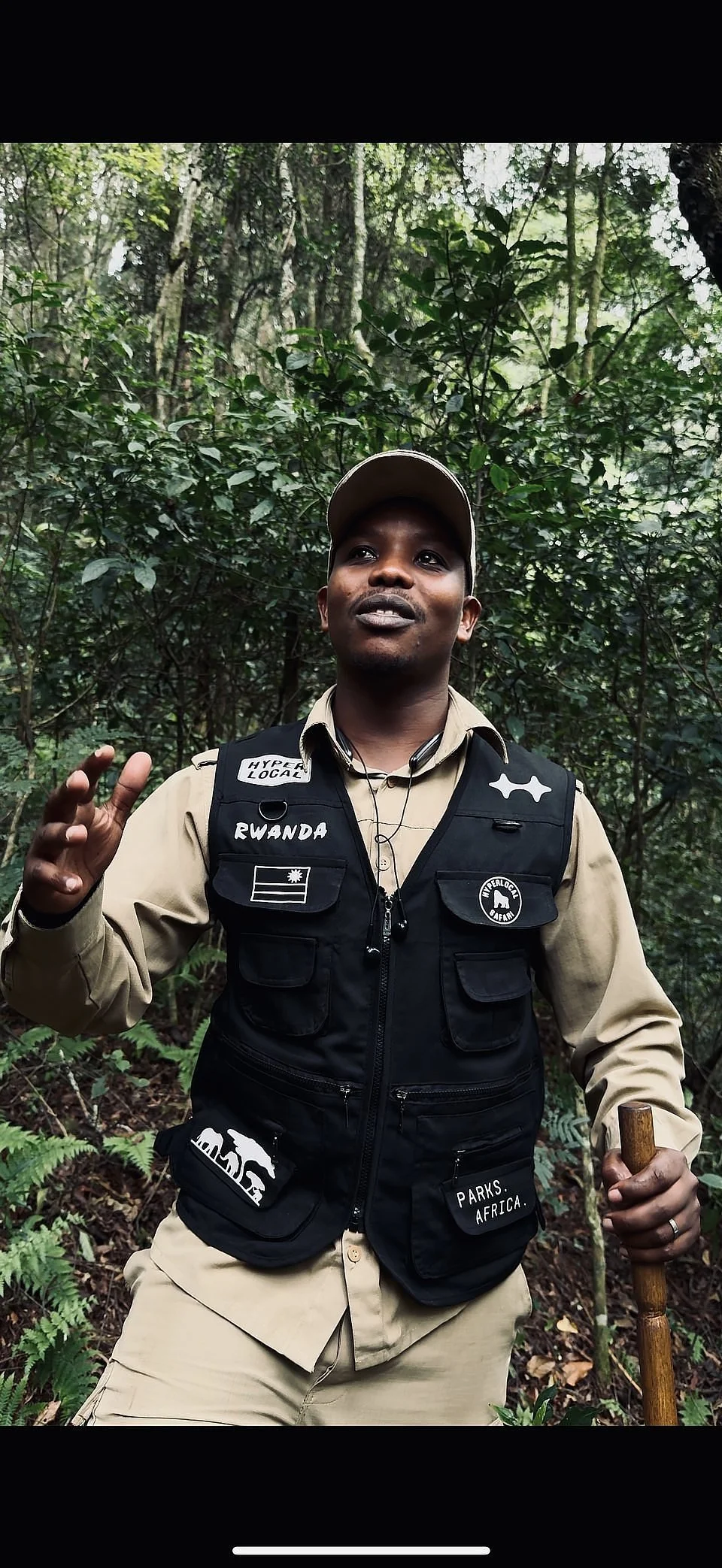 A wildlife ranger in a tan uniform and black vest with patches and text, standing in a forest with dense green foliage, holding a wooden walking stick and gesturing with his hand.