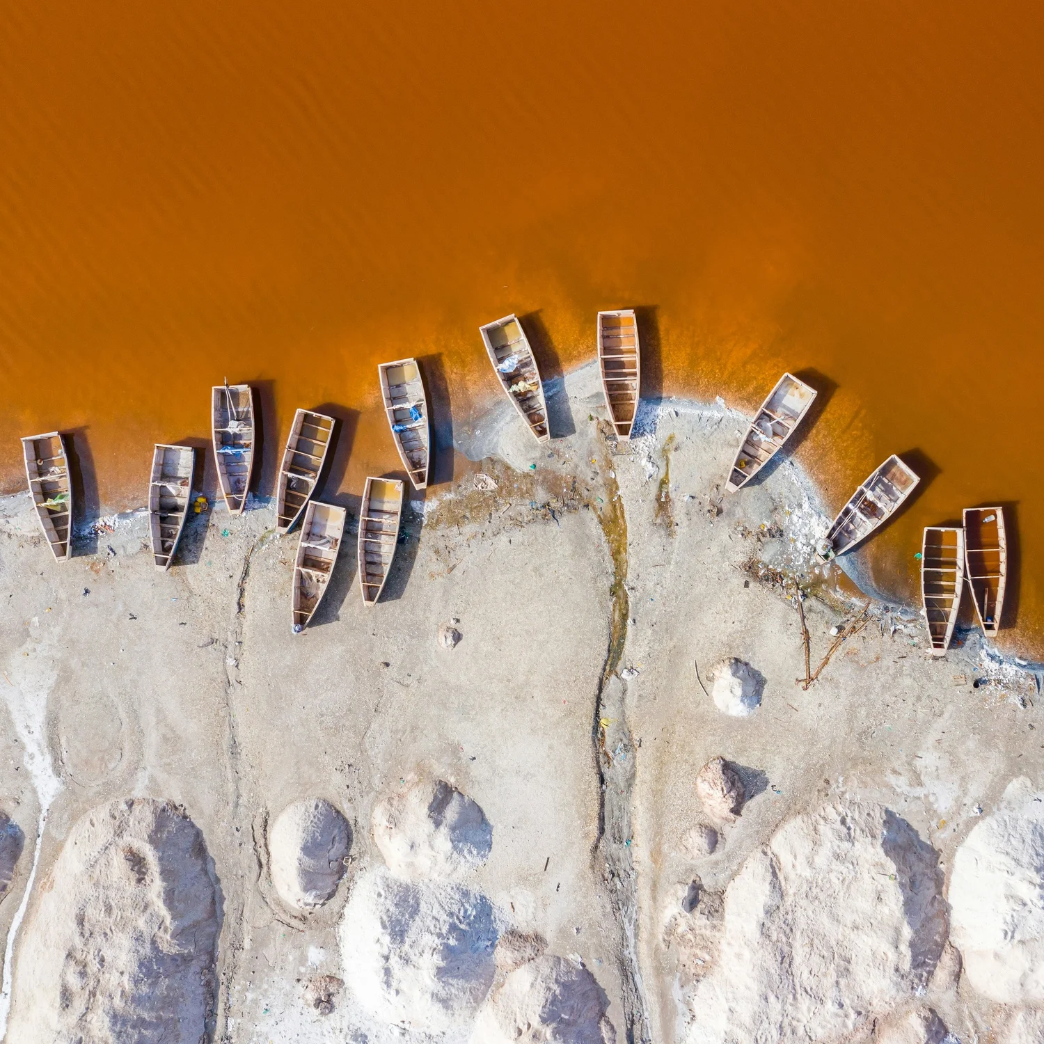 Aerial view of wooden boats lined up along a sandy shoreline with orange-colored water in the background.