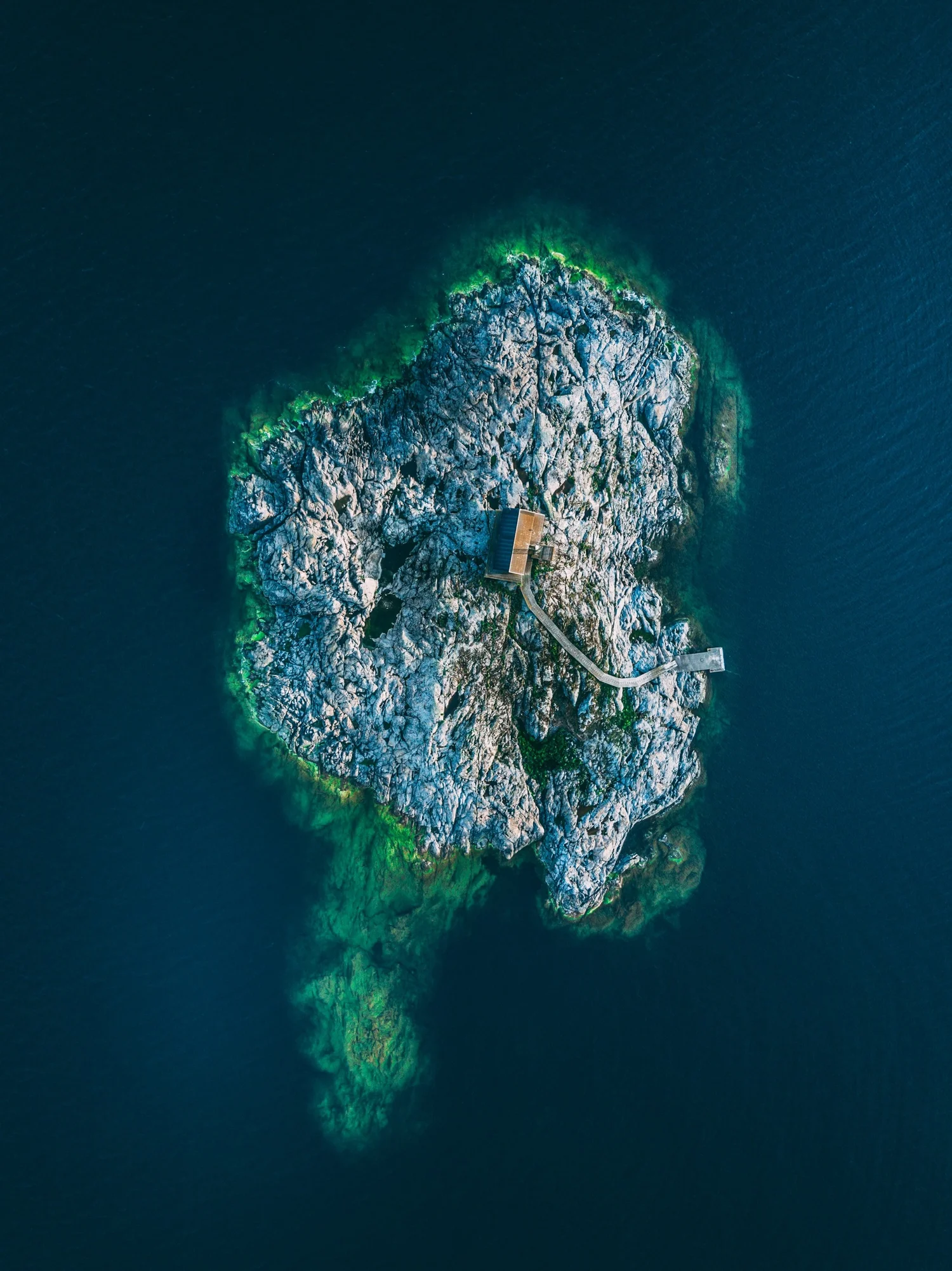 An aerial view of a rocky small island surrounded by deep blue water, with a walkway connecting to a small building on the island.