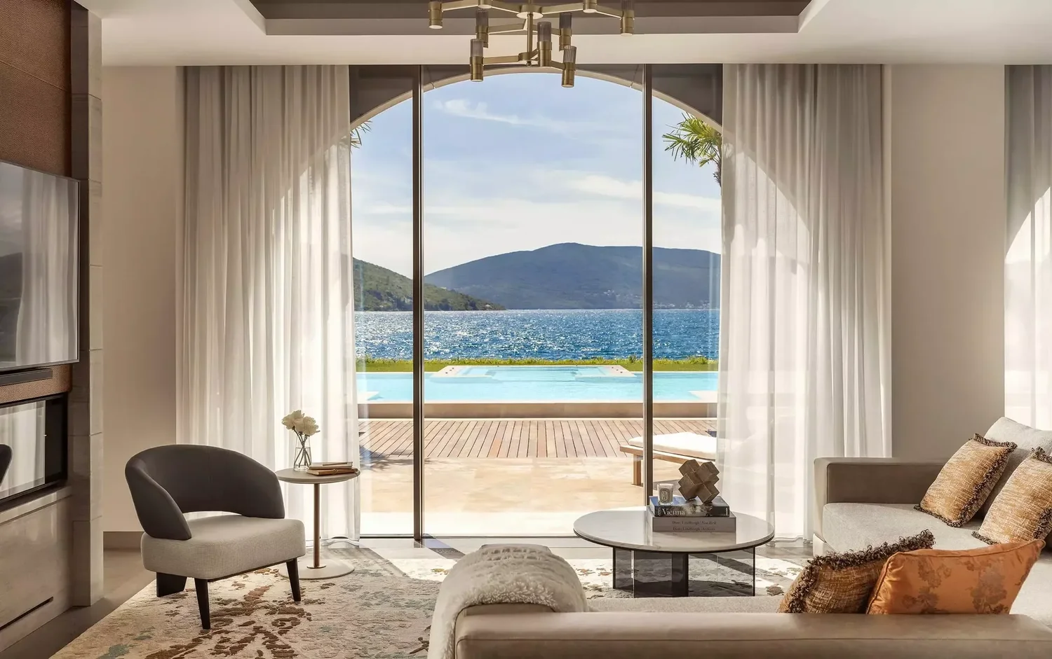 Living room with large glass sliding doors opening to a view of a pool, lake, and mountains in the distance, with white curtains and modern furniture.