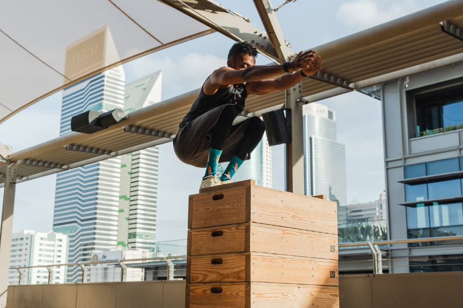 Young man jumping onto a wooden box in an outdoor urban gym with city buildings in the background.