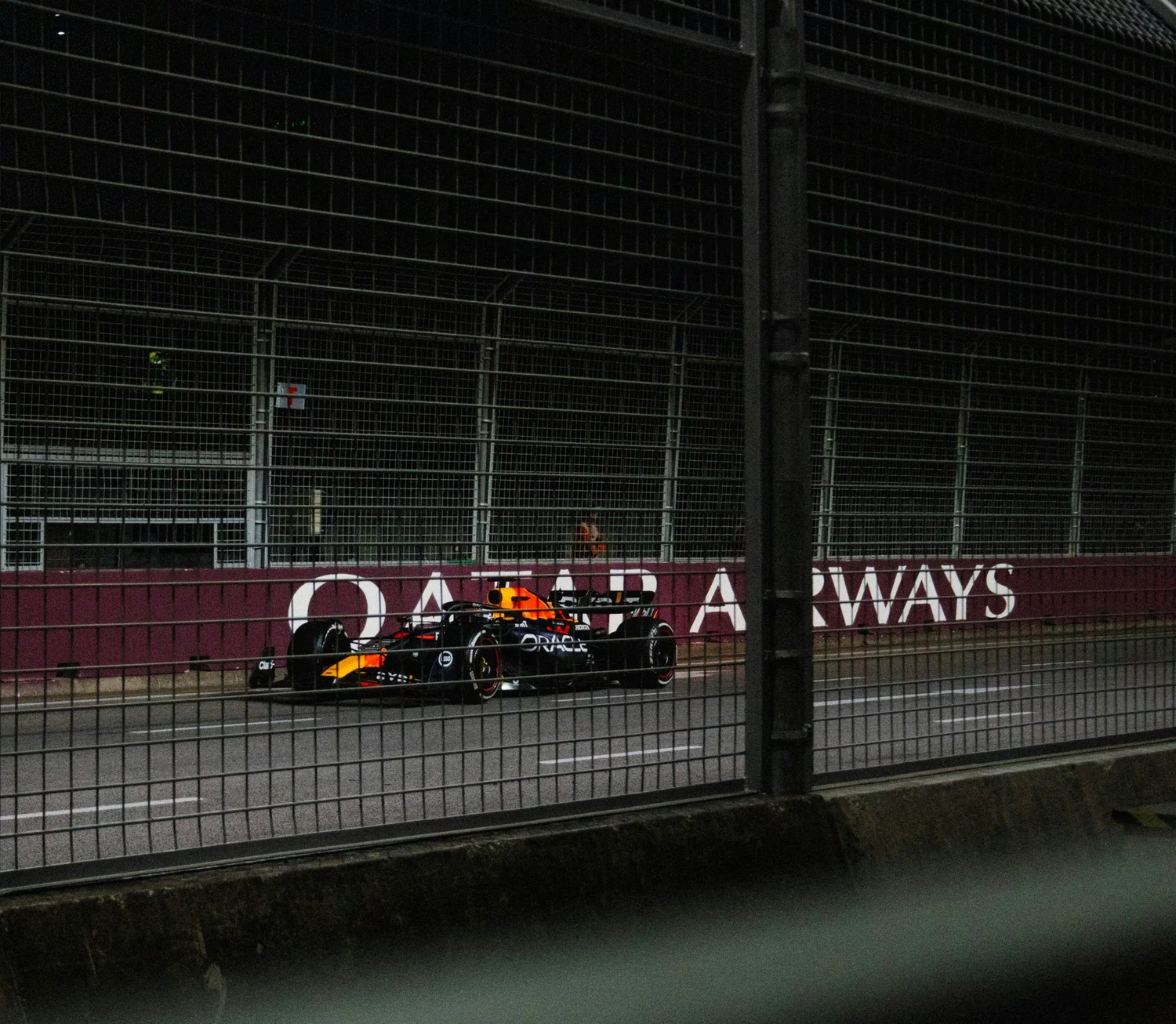 A Formula 1 race car driving on a track behind a metal fence at night, with signage that reads 'QATAR ALWAYS' in the background.