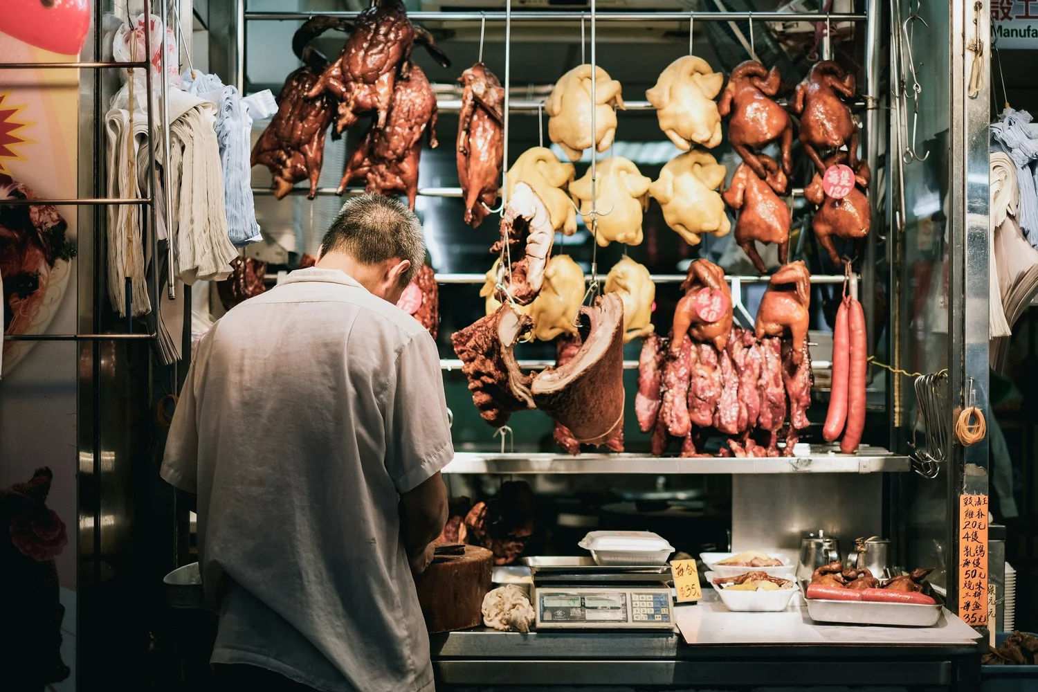 A street market stall displaying hanging roasted poultry, ducks, and sausages. A man is preparing food behind the counter.
