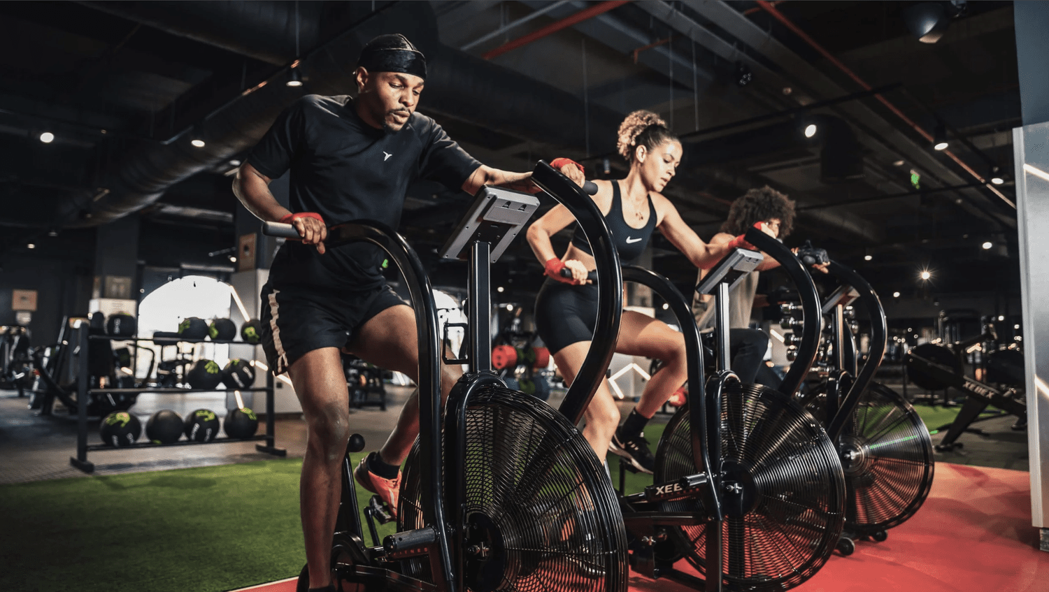 Three people working out on air resistance exercise bikes in a gym, with gym equipment and weight balls in the background.