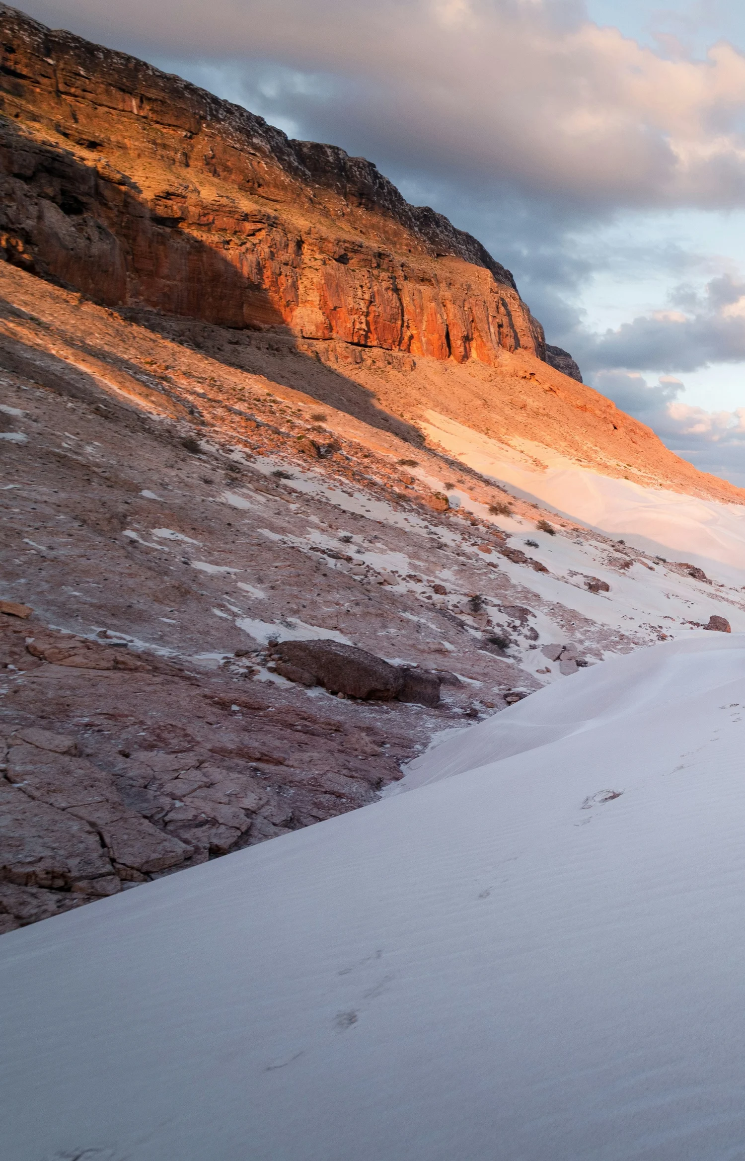 Snow-covered slope with rocky, reddish-brown cliffs in the background under a partly cloudy sky during sunset.