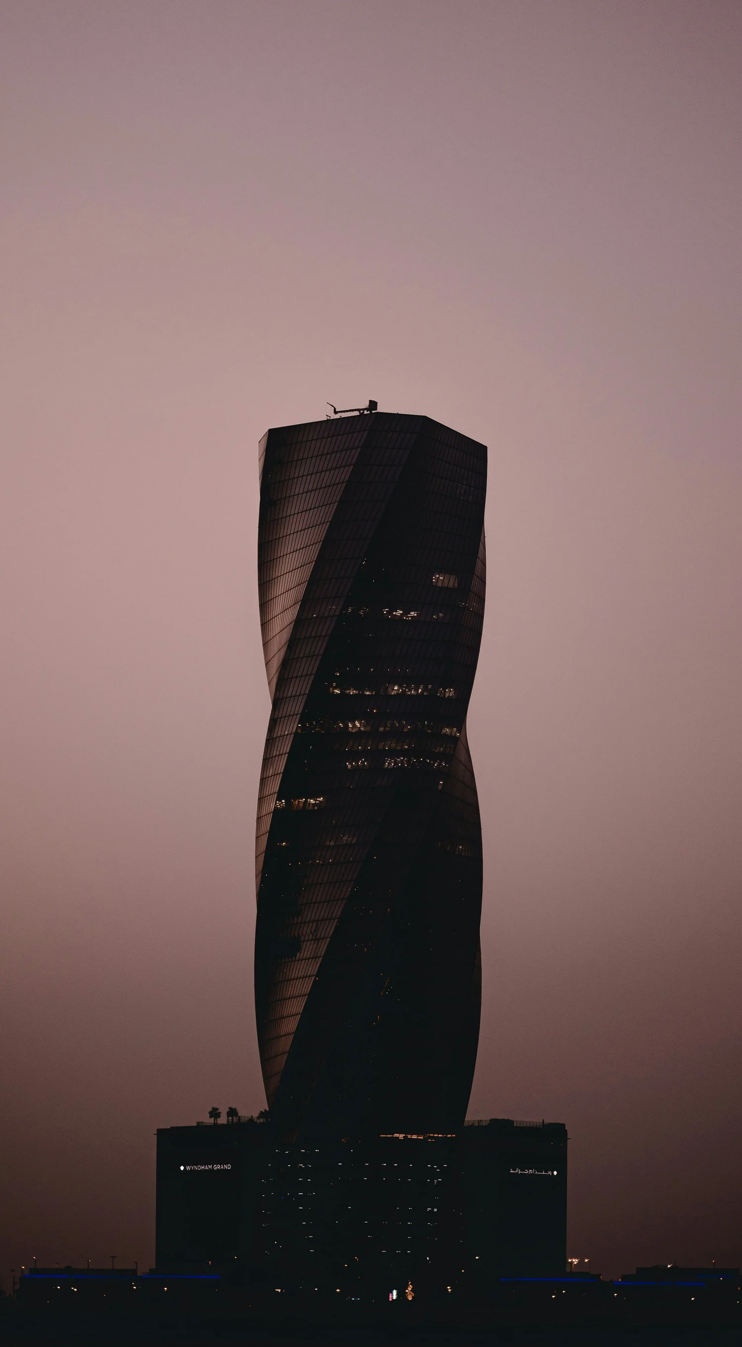A tall, modern skyscraper with a twisted design against a dusky sky, with some lights visible inside and on the building.