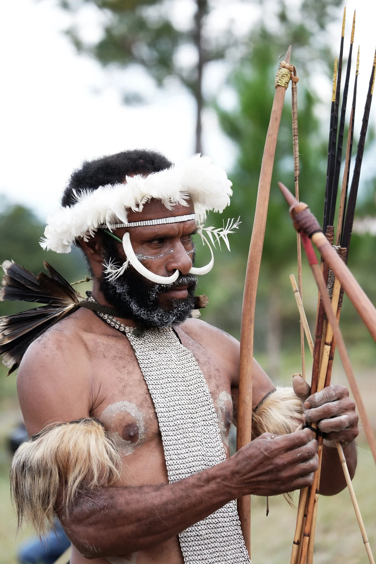 A man dressed in traditional tribal attire with face paint, wearing a feathered headband and jewelry, holding arrows, and standing outdoors.