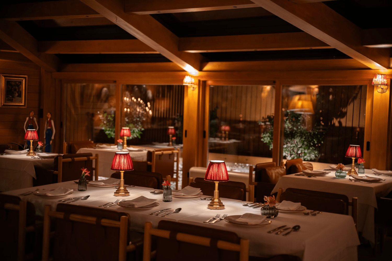 Elegant restaurant interior with wooden walls, large windows, and red-lamped lamps on the tables, set for dining with white tablecloths, plates, silverware, and small floral centerpieces.