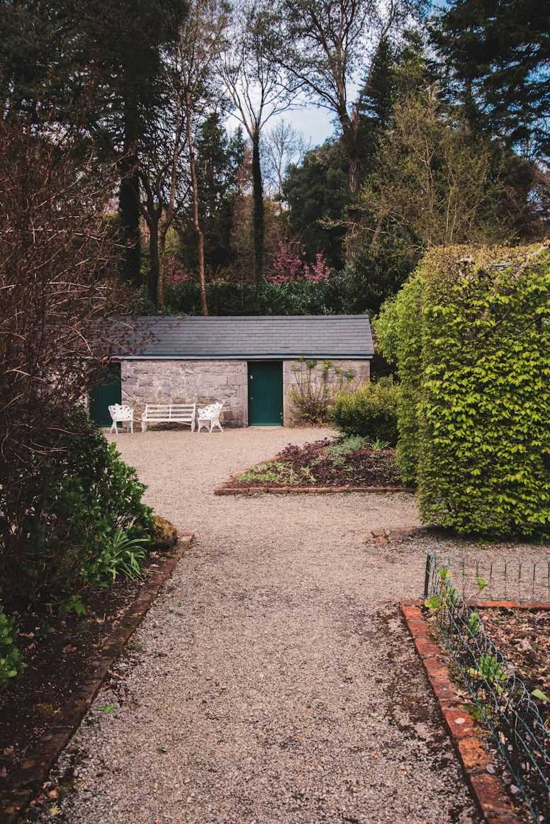 A garden path leading to a stone shed with a grey roof, flanked by green bushes and trees, with two white benches in front of the shed and a small garden bed on the right.