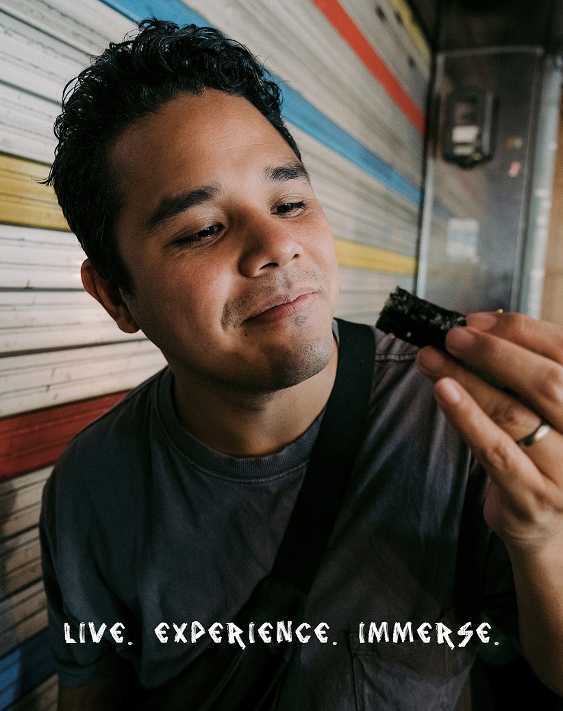 A man with dark curly hair looks at a piece of sushi he is holding in his right hand, with a colorful wooden wall in the background. Hyperlocal World of Travel.