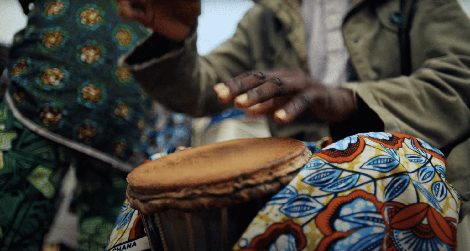 Two people playing a drum, with one person's hand striking the drum, and both wearing colorful, patterned clothing.