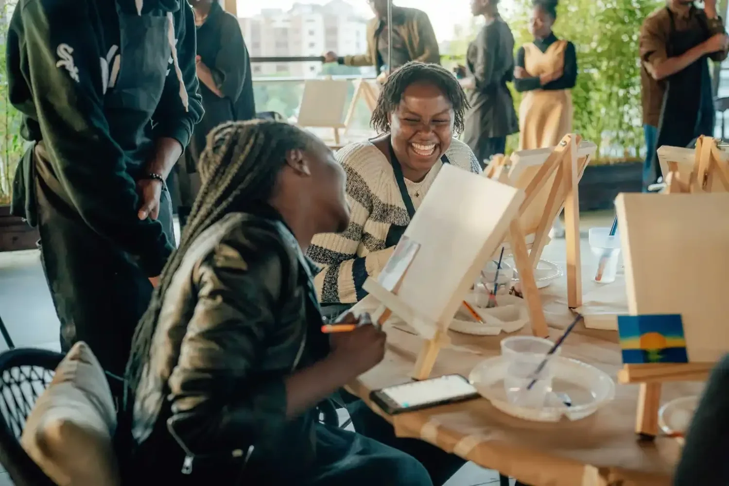 People participating in an art workshop, with two women painting on canvases on easels, smiling and engaging in conversation.