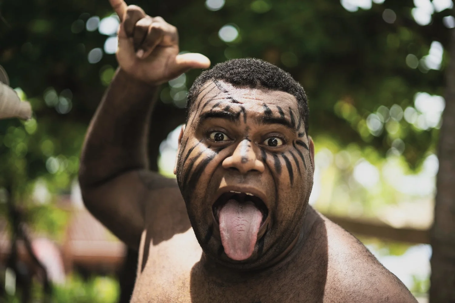 A man with face paint making a wild face, sticking out his tongue, with trees and sunlight in the background.