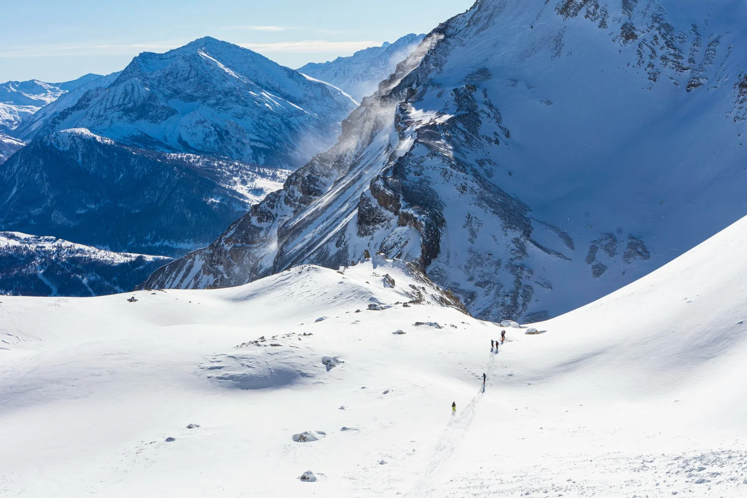 Snow-covered mountain landscape with a group of climbers or skiers traversing the snow in the foreground and rugged peaks in the background.