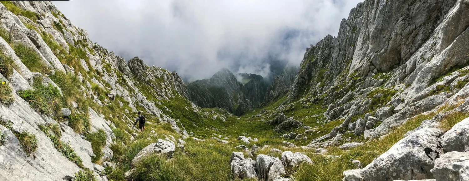 Hiker walking through a rocky mountain valley with steep cliffs on either side, partly shrouded in clouds.