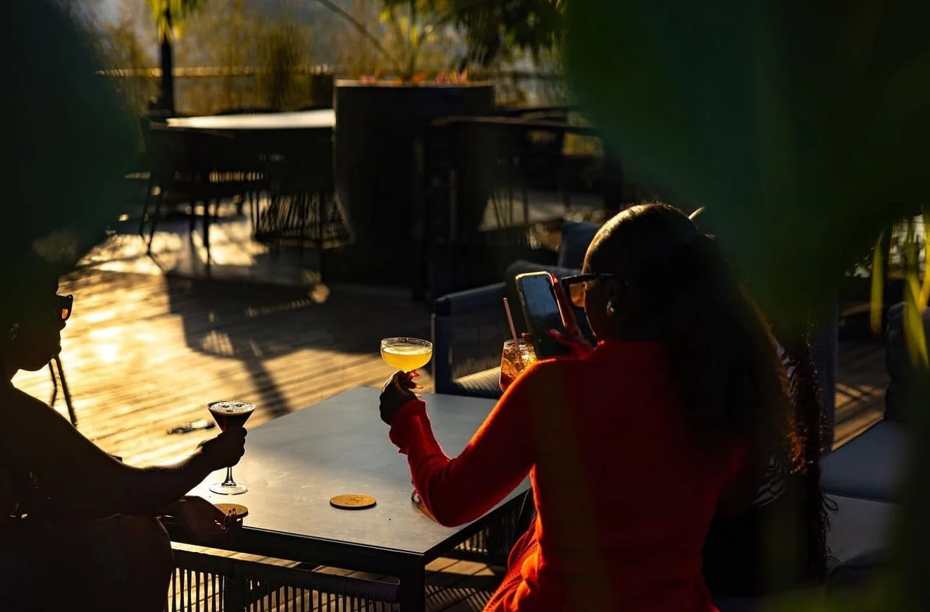 Two women sitting at an outdoor patio table on a sunny day, one holding a cocktail and the other taking a photo with a smartphone, with shadows and trees in the background.