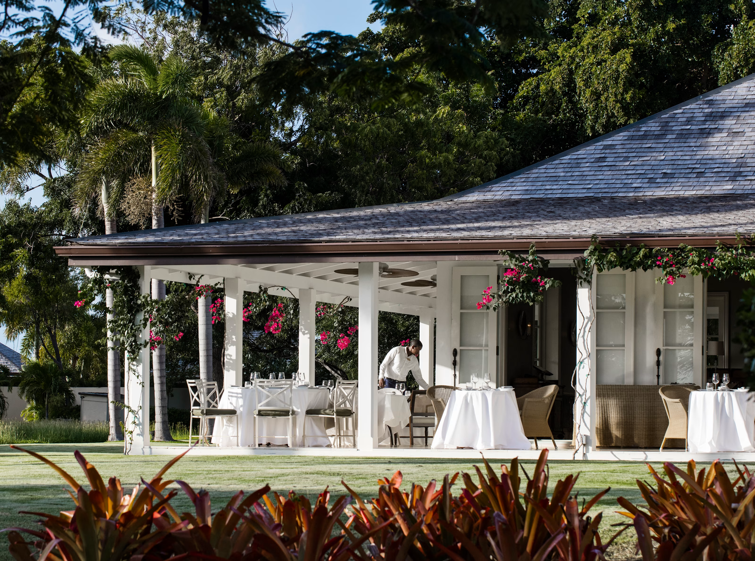 Outdoor dining area with white tablecloth-covered tables and chairs on a patio, decorated with pink flowers and surrounded by green plants and trees, with a person setting the table.