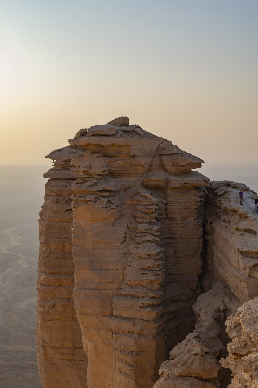 A large sandstone cliff with layered, weathered rocks near a flat, sandy landscape. A few small people are visible on the right side of the cliff for scale, with a clear sky in the background at sunset or sunrise.