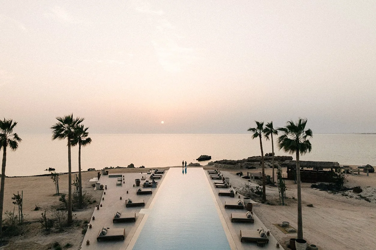A serene beach scene at sunset with a long pool leading to the ocean, surrounded by lounge chairs, palm trees, and rocks.