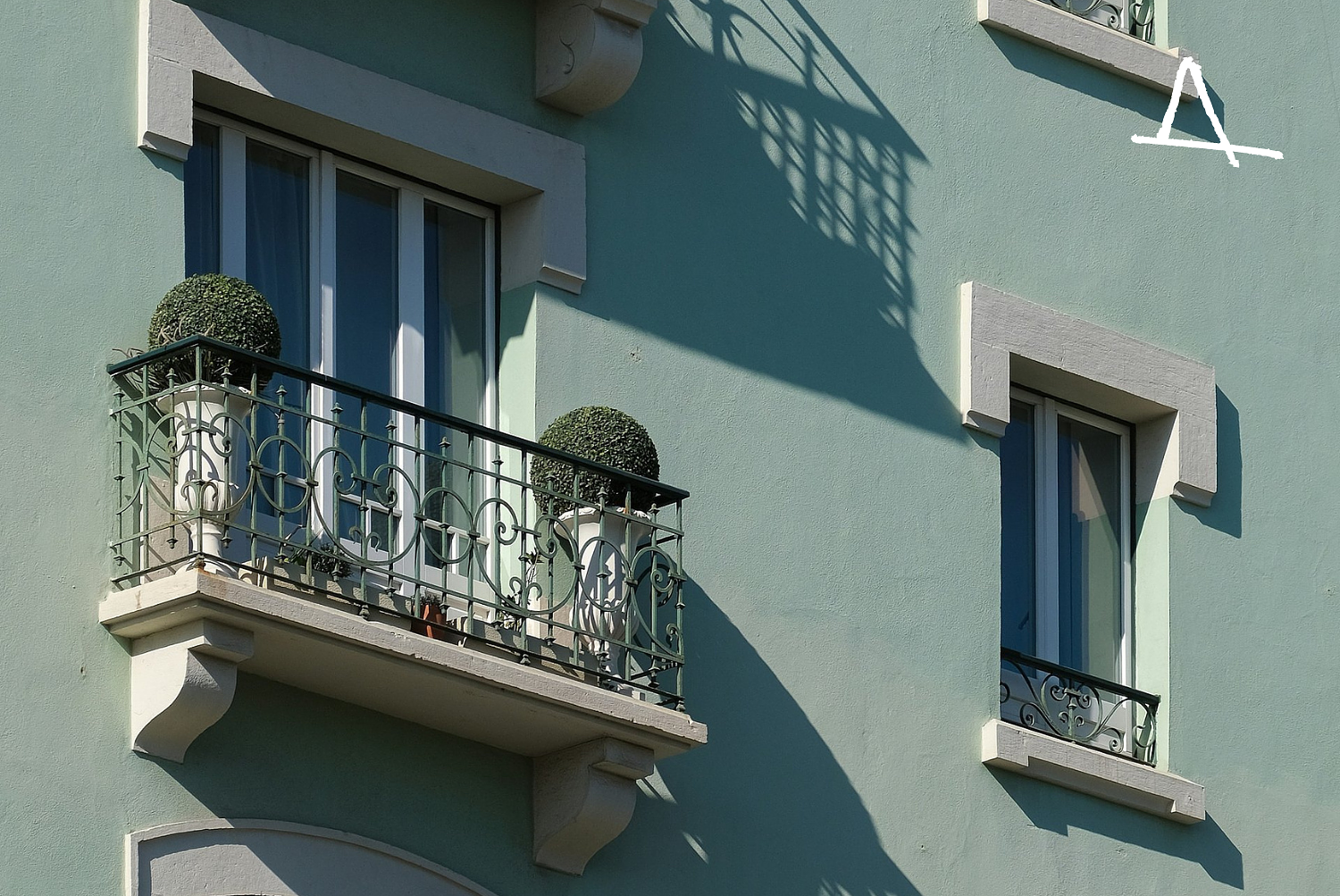Close-up of a light green building with two windows, one with a small balcony and potted plants, and decorative white trim. A shadow of a balcony overhang is cast on the wall.